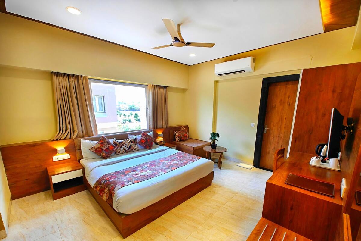 A hotel room with a large bed, bedside tables, a desk, a chair, a ceiling fan, an air conditioning unit, and a window with curtains.