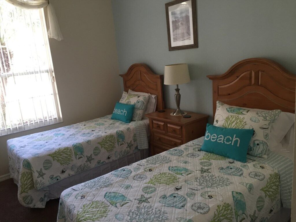 Ein Schlafzimmer mit zwei Betten, jeweils mit einem an den Strand erinnernden Bettbezug, einem Nachttisch mit Lampe und einem Fenster mit Jalousien.