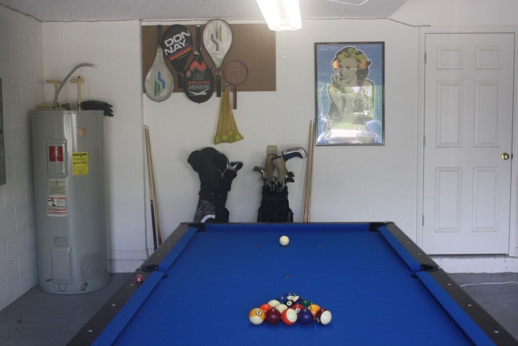 A pool table with balls, a water heater, and sports equipment against a white wall.
