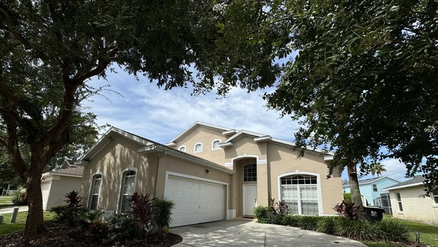 A single-story house with a beige exterior, a white garage door, and a front yard with a tree and shrubs. - Julliard Villa (Davenport)