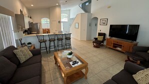 A modern living room with a sofa, coffee table, TV, and a kitchen area with bar stools. - Julliard Villa (Davenport)