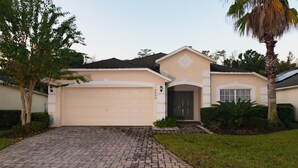 A two-story house with a beige facade, a garage, and a front yard with palm trees and shrubs. - Cumbrian Retreat (Kissimmee)