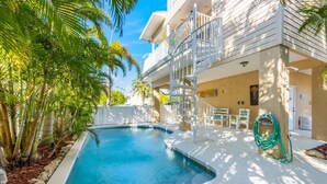 A swimming pool with a white deck, surrounded by palm trees and a building with a balcony. - Gigi & Poppy’s Paradise: Pool, Gulf Views, Walk to Beach (Holmes Beach)