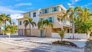 A two-story house with a front porch, a garage, and a small front yard with palm trees. - Gigi & Poppy’s Paradise: Pool, Gulf Views, Walk to Beach (Holmes Beach)