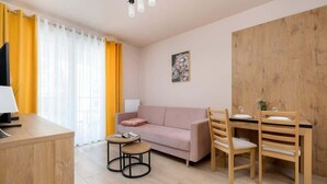 A modern living room with a sofa, a wooden dining table with chairs, a small round table with a lamp, and a painting on the wall.