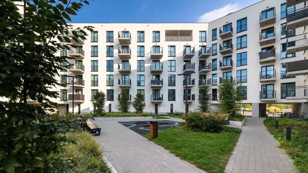 A modern apartment building with multiple balconies, a central courtyard with a circular seating area, and surrounding greenery.
