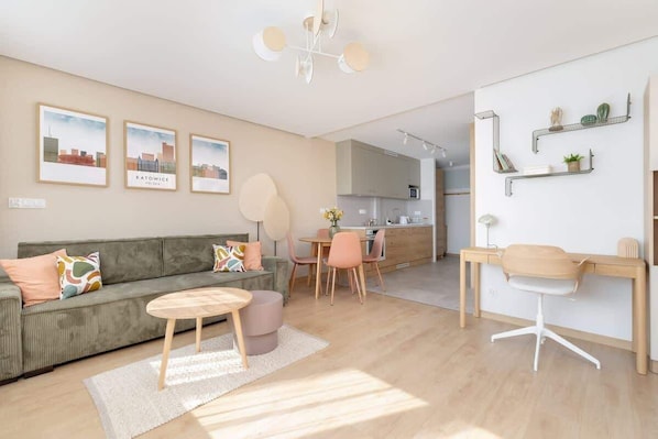 Un salon moderne avec un canapé, une table basse et un coin repas. On peut voir des tableaux encadrés au mur, une cuisine à l’arrière-plan et un bureau avec une chaise.