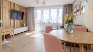 A modern living room with a wooden panel wall, a flat-screen TV, a round dining table with pink chairs, and a vase with flowers. - Stylish Apartment in the Center | Air Conditioning, Balcony (Katowice)