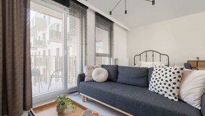 Ein modernes Wohnzimmer mit einem grauen Sofa, einem Holztisch mit Gebäck, einem Bett mit schwarzem Gestell und Blick auf ein Gebäude.