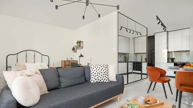 Ein modernes Wohnzimmer mit grauem Sofa, einem Esstisch mit orangefarbenen Stühlen, einer Küchenzeile und einem großen Fenster.