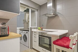 A compact kitchen with a washing machine, a toaster, a small countertop, and a red chair. - Pau - Habitat Apartments (Barcelona)