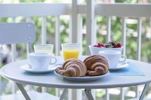 Mesa branca com croissants, uma tigela de frutas e dois copos de suco de laranja para o café da manhã.