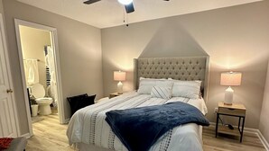 Un dormitorio con una cama, dos mesitas de noche con lámparas, un ventilador de techo y una puerta que da a un baño.