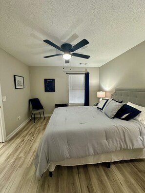 A bedroom with a bed, a ceiling fan, a window with blinds, a chair, and a bedside table with a lamp.