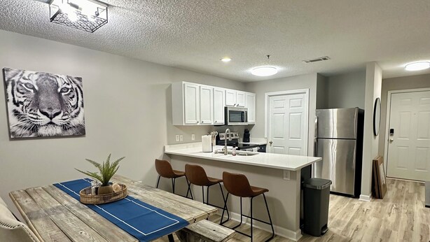 Una cocina moderna con armarios blancos, un refrigerador de acero inoxidable y un comedor con mesa y sillas de madera.