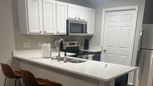 Una cocina moderna con armarios blancos, un refrigerador de acero inoxidable, un microondas y un fregadero. Hay tres taburetes de bar y un cubo de basura.