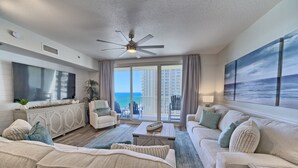 A spacious living room with a large flat-screen TV, a ceiling fan, and a view of the ocean through large sliding doors.
