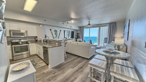 A modern kitchen with built-in appliances, a dining area with a round table and benches, and a balcony with a view of the ocean.