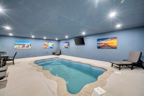 Una piscina interior con borde ondulado, rodeada de sillas de descanso y un televisor de pantalla plana montado en la pared.