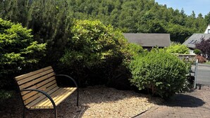 A bench in a landscaped area with a paved path, surrounded by dense greenery and a hill in the background.
