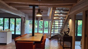 Un vaste espace dédié à la salle à manger, avec une table en bois et des chaises orange, de grandes fenêtres offrant une vue sur l’extérieur, et un escalier menant à un niveau supérieur.