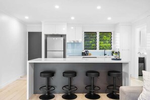 A modern kitchen with a central island, bar stools, a refrigerator, and a window with greenery.