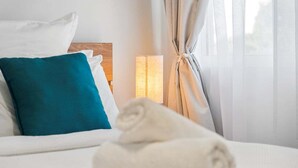 Una cama bien hecha con almohadas blancas y azul verdosas, una toalla blanca enrollada y una lámpara de noche.