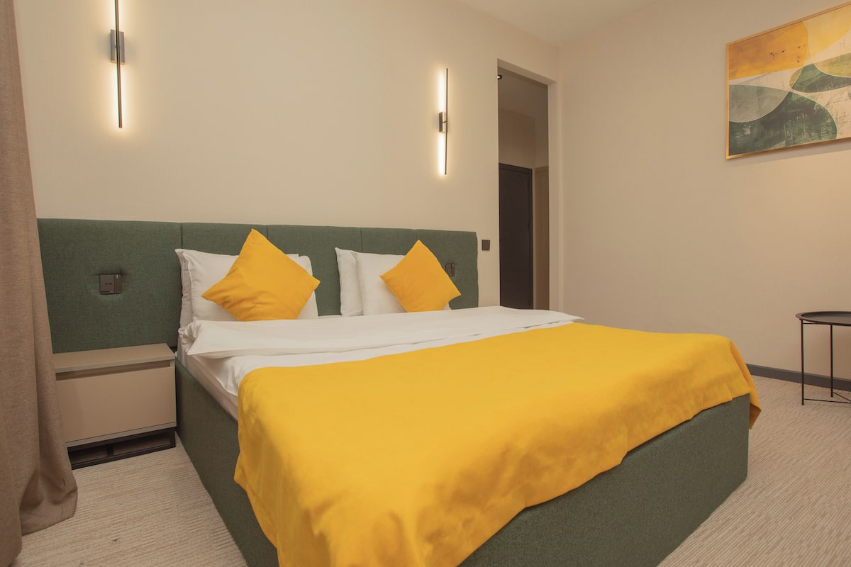 A hotel room with a bed, green headboard, white and yellow pillows, a yellow bedspread, a nightstand, and a painting on the wall.