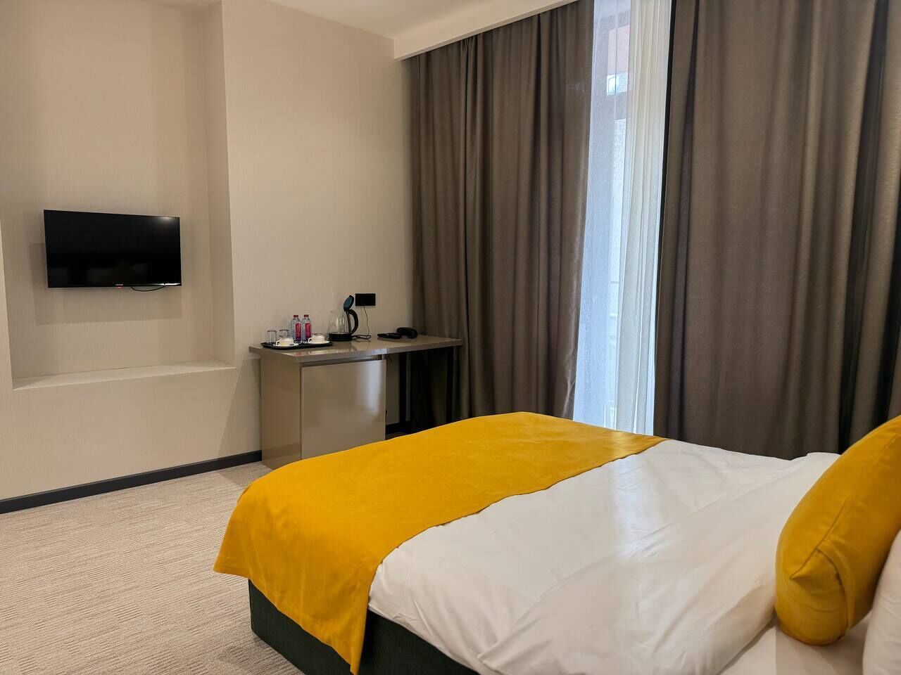 A hotel room with a bed, a desk, a flat-screen TV, and a window with curtains.