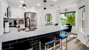A modern kitchen with white cabinets, a stainless steel refrigerator, a large island with bar stools, pendant lighting, and a view of greenery through large windows. - Beautifully Updated 2 Bedroom Condo with Stunning Views (La Quinta)