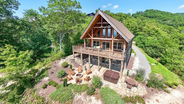 A wooden cabin with a balcony, surrounded by lush greenery and a stone patio with outdoor seating. - Altitude Adjustment Magnificent Custom Cabin with Breathtaking Views (Bryson City)
