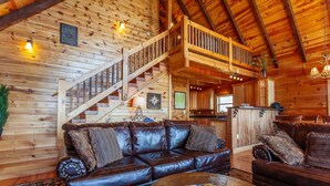 A cozy log cabin interior with a leather sofa, a coffee table, and a staircase leading to a loft area. - Altitude Adjustment Magnificent Custom Cabin with Breathtaking Views (Bryson City)
