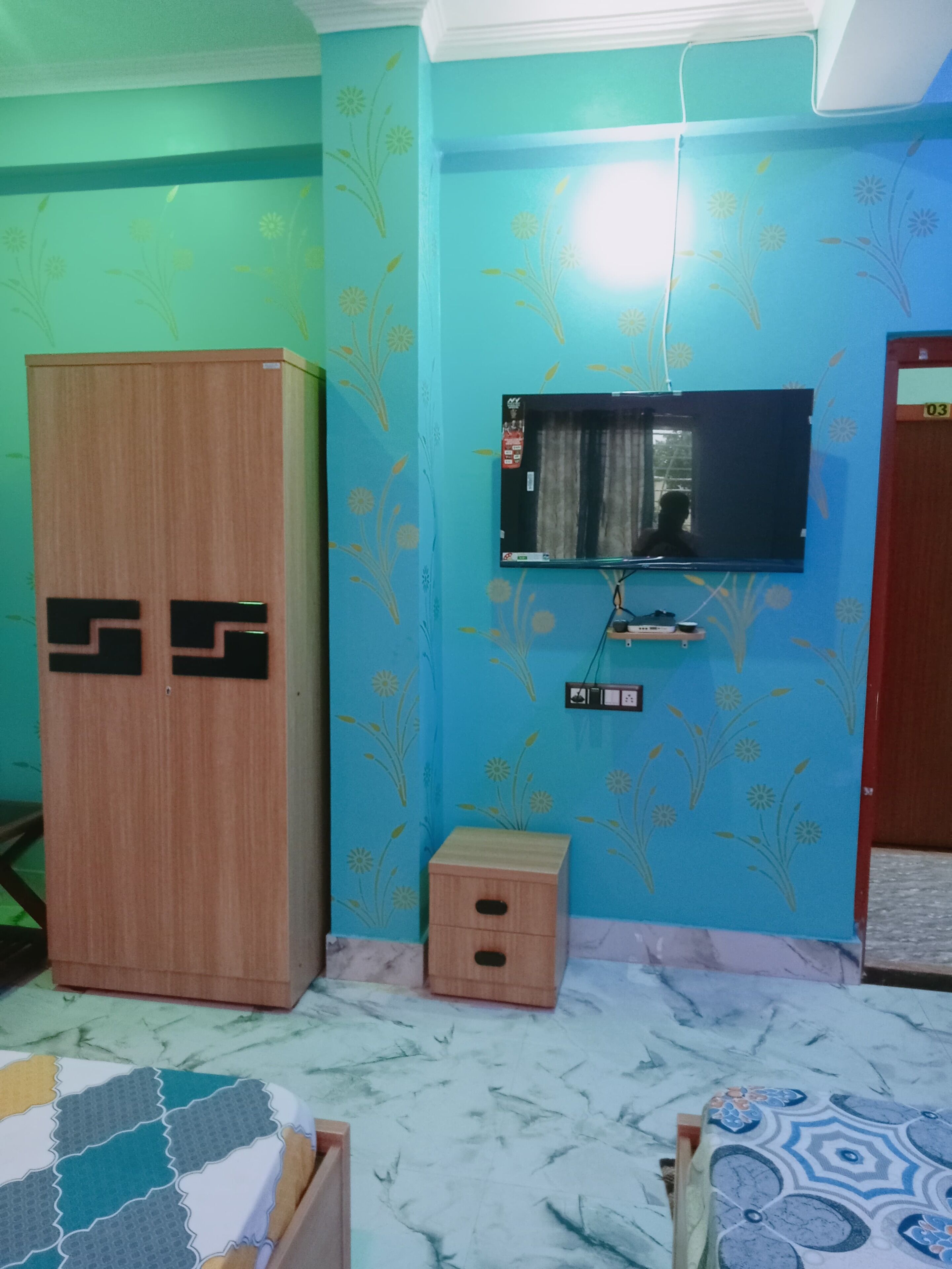 Una habitación con un armario de madera, un televisor montado en la pared, un pequeño mueble de madera y una colcha estampada.