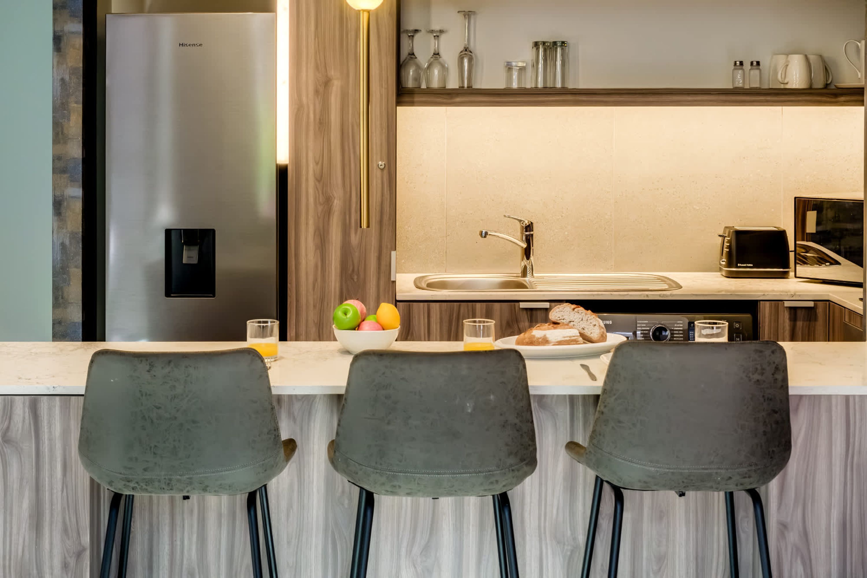 A modern kitchen with a stainless steel refrigerator, a sink, and a toaster. There are three chairs in front of the counter.