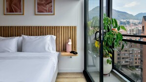 A modern bedroom with a wooden headboard, a bedside table with a lamp, a large window with a view of a cityscape and mountains, and two framed artworks on the wall. - w Amazing Loft w Balcony in Chapinero (Bogotá)