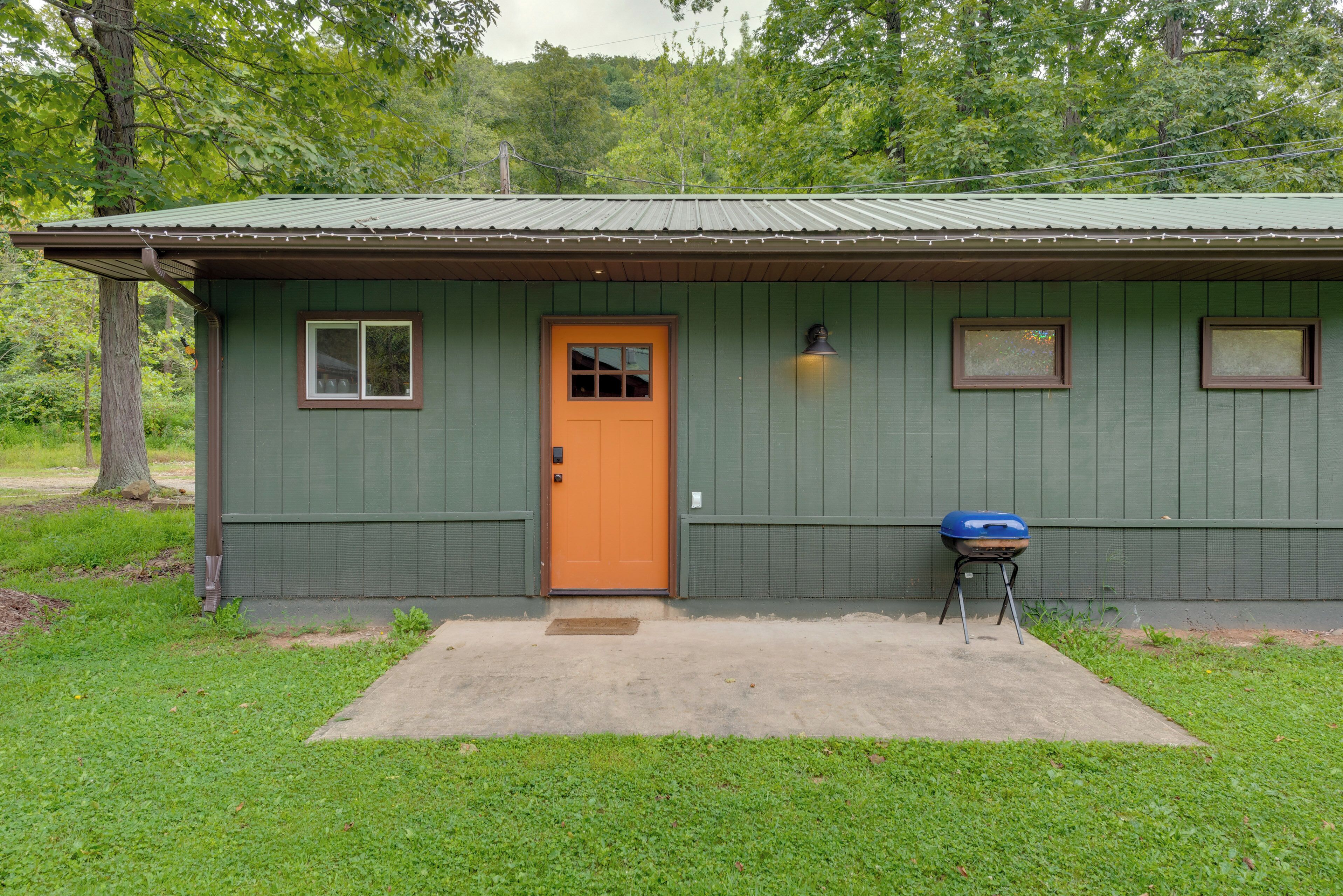 A small, single-story building with a green exterior and a brown door. There is a small porch area with a blue grill and a single window on the side.