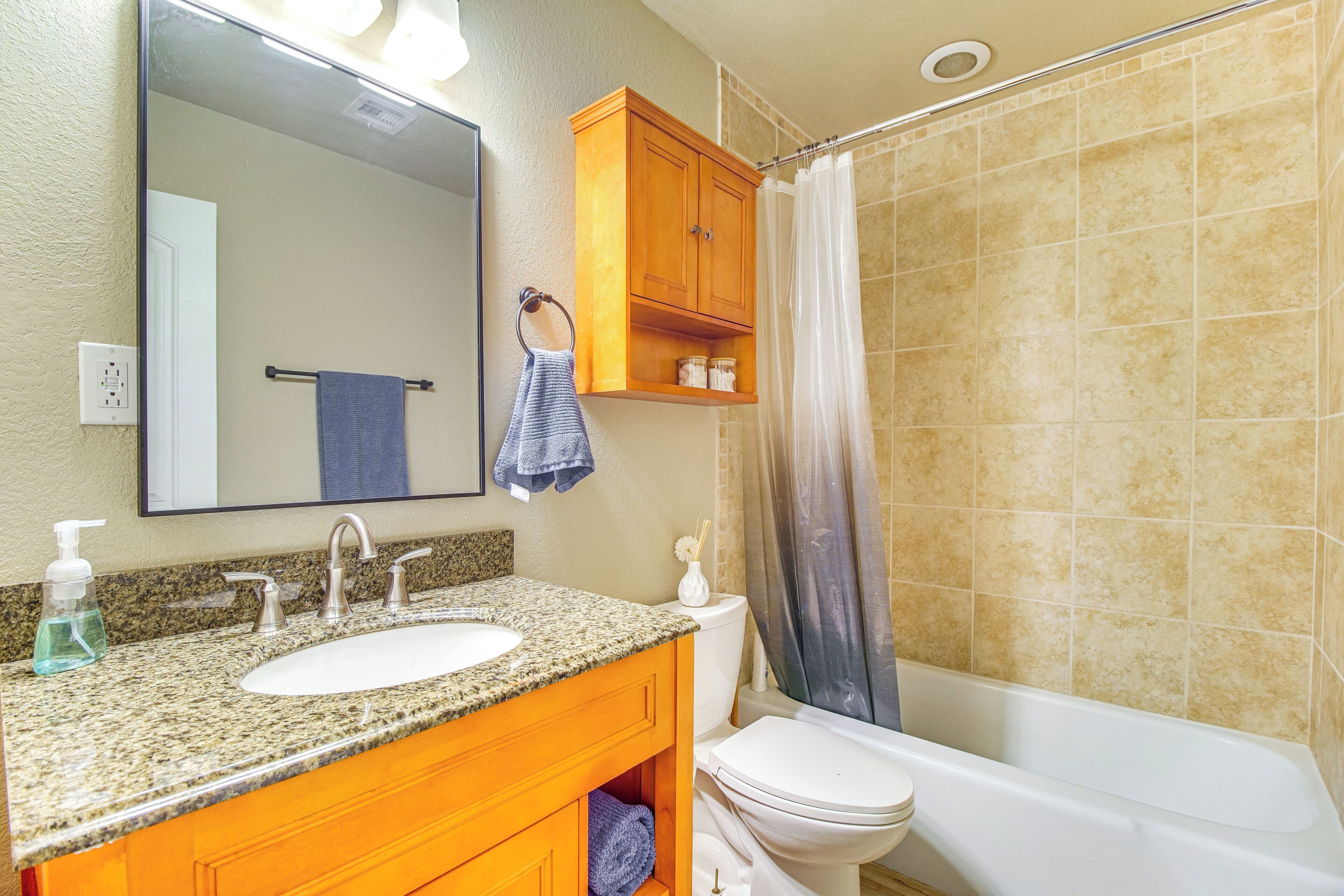 A bathroom with a granite countertop sink, wooden cabinets, a white toilet, and a shower with a beige tiled wall.