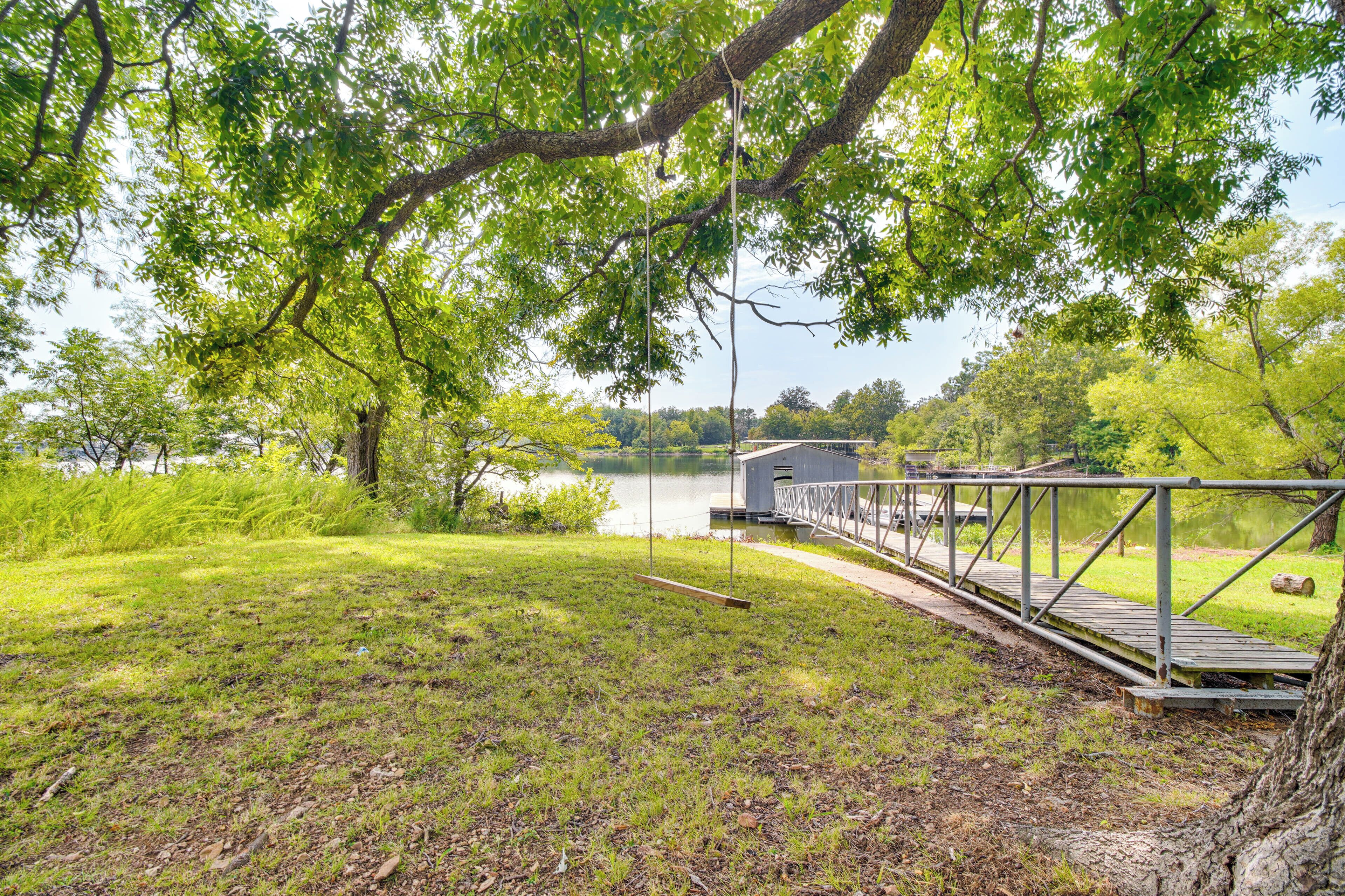 A wooden dock extending over a calm lake, surrounded by lush greenery and a small building with a metal railing.