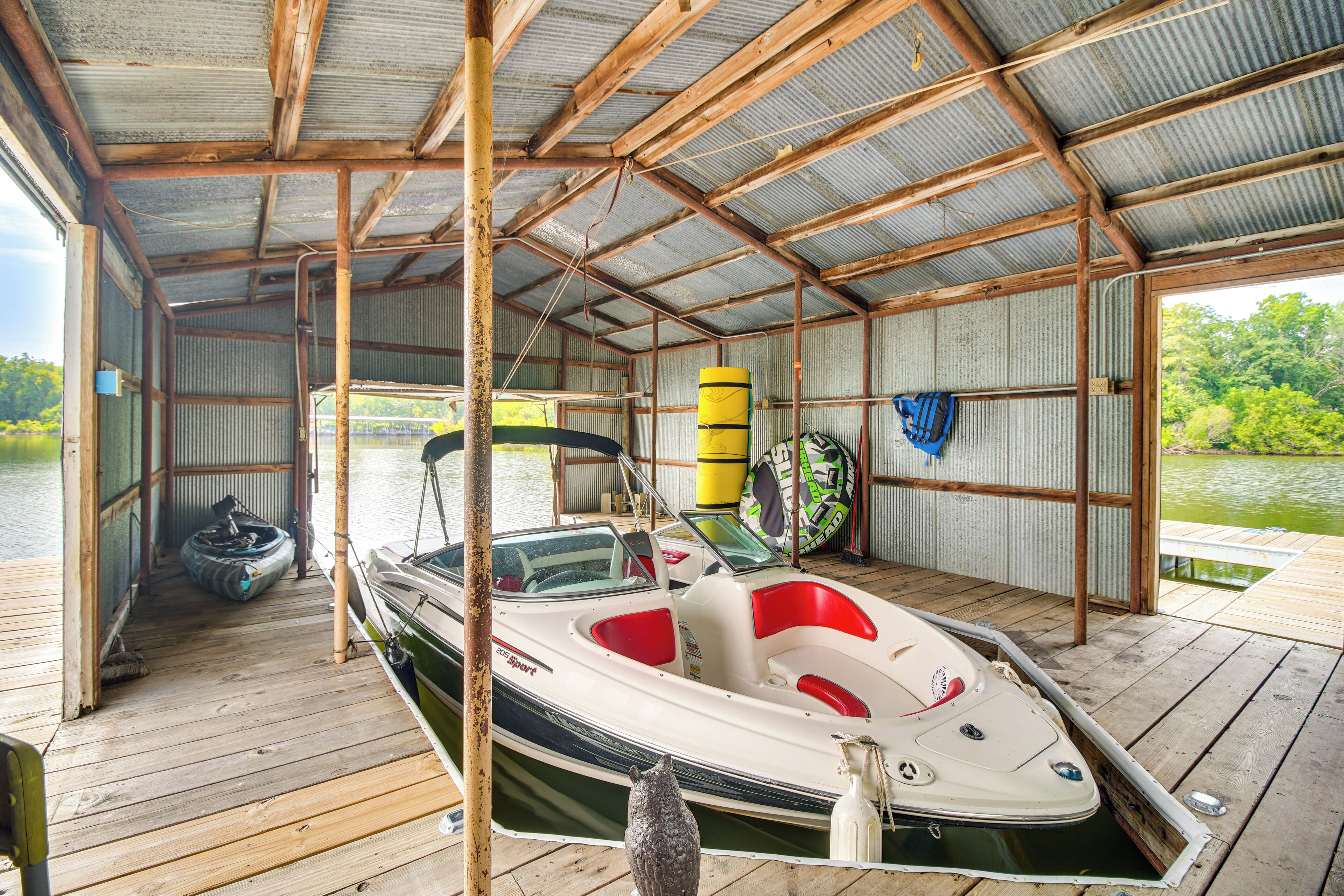 A covered boat dock with a wooden floor, a boat with red and white seating, and a kayak and paddleboard stored against the wall.