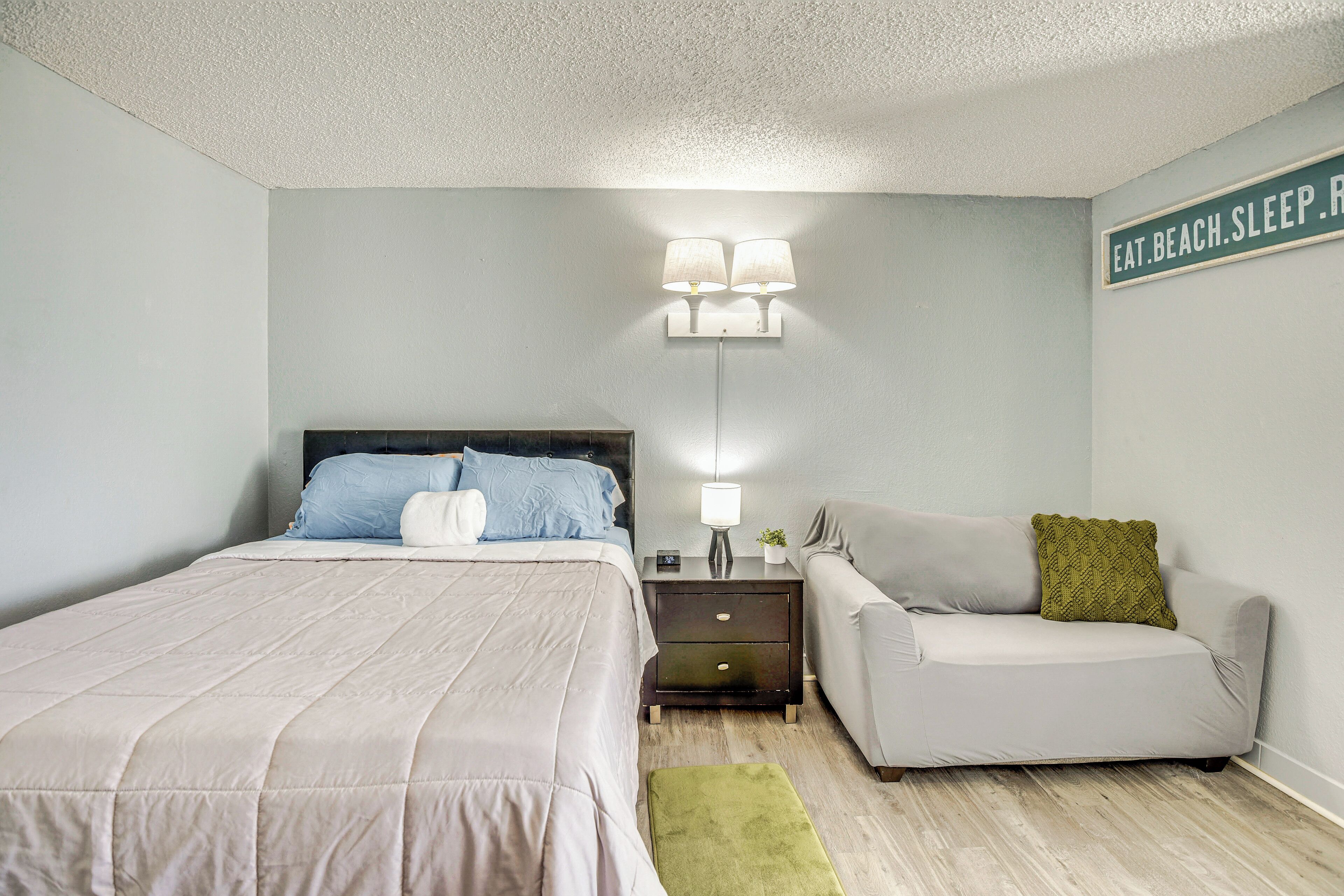 A bedroom with a bed, a nightstand, a lamp, a sofa, and a sign on the wall.
