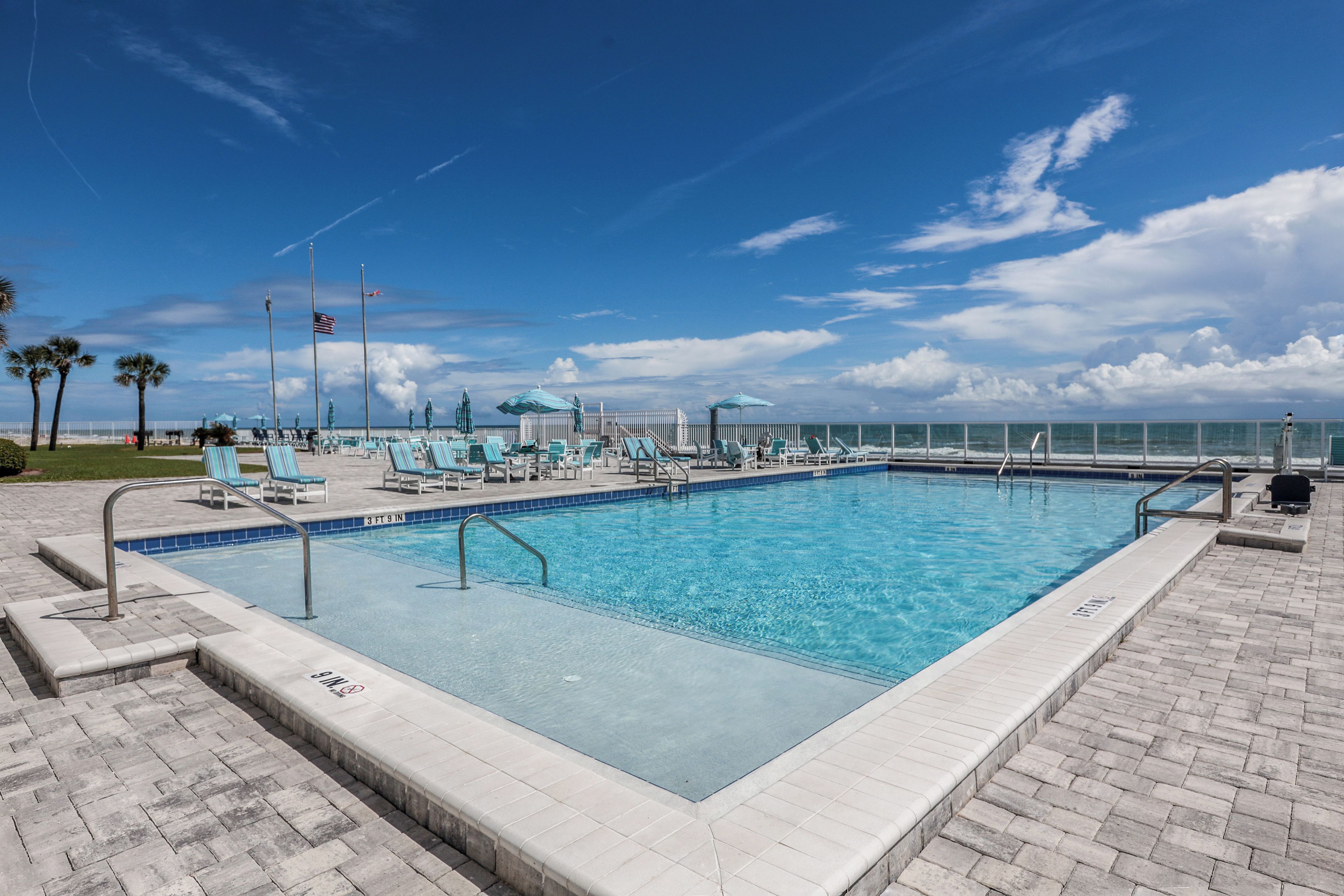 Una piscina rectangular con bordeado de azulejos, rodeada de sillas de descanso y sombrillas, con vista despejada al océano y un asta de banderas.