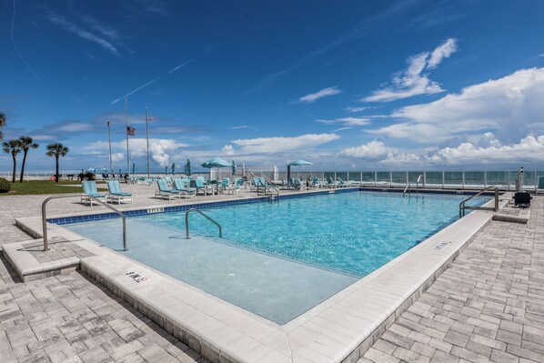 A rectangular swimming pool with a tiled edge, surrounded by lounge chairs and umbrellas, with a clear view of the ocean and a flagpole with flags.