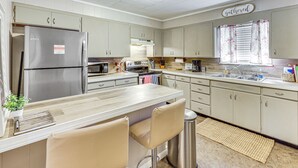 Eine Küche mit einem Edelstahl-Kühlschrank, einer Mikrowelle, einem Herd und einem Spülbecken. Zwei Barhocker und ein kleiner Mülleimer sind ebenfalls vorhanden.