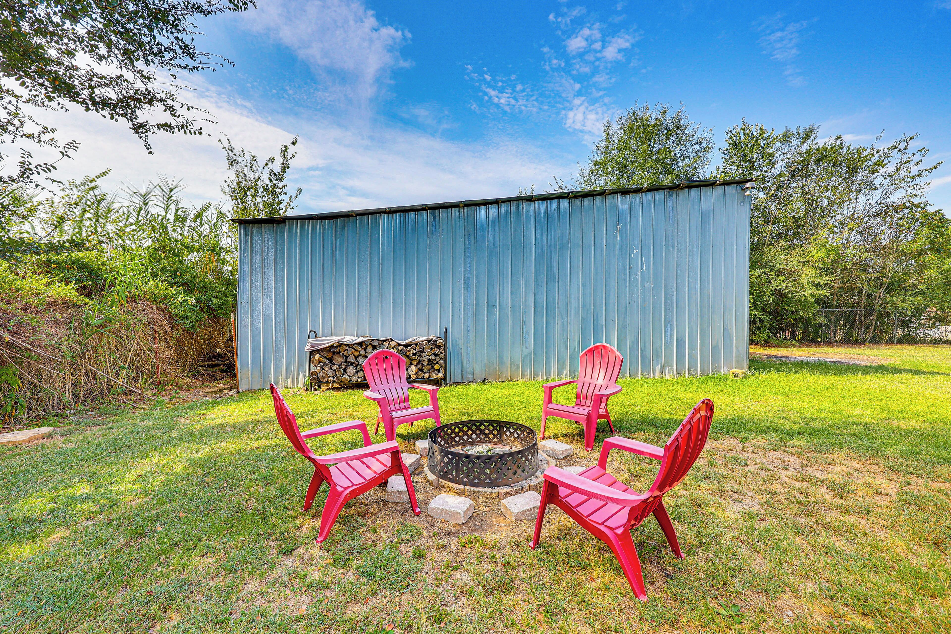 A backyard with a fire pit surrounded by four red Adirondack chairs, a metal shed, and a stack of firewood.