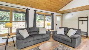 Ein gemütliches Wohnzimmer mit zwei grauen Sofas, einem Holztisch und großen Fenstern mit Blick auf den Wald.