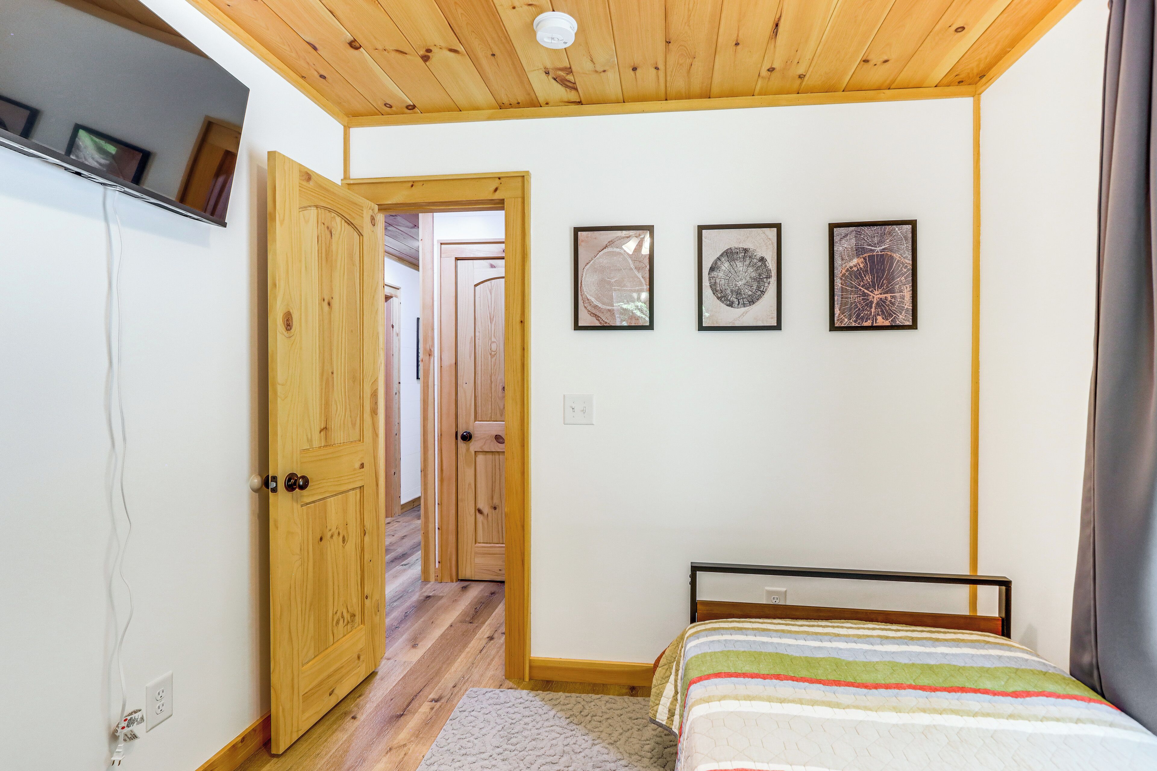 A bedroom with a wooden ceiling, a bed with a striped bedspread, and three framed artworks on the wall.