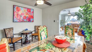 A dining area with a glass table, chairs with patterned upholstery, a ceiling fan, and a colorful abstract painting on the wall.