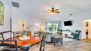 A spacious living room with a wooden dining table, comfortable seating, a ceiling fan, and a large window providing natural light.