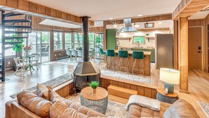 Een ruime woonkamer met een grote open haard, comfortabele zithoek en een zichtbare keuken achter een open deur.