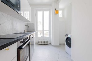 A kitchen with a black countertop, white cabinets, and a built-in oven. There is a window with white frames and a radiator below it. A washing machine is visible in the corner. - Résidence Neuilly BLOUD (Neuilly-sur-Seine)